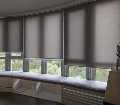 Motorized roller shades in the interior. Automatic roller blinds beige color on big glass windows. Home luxury curtaines are above the windosill with pillows. Summer. Green trees outside.